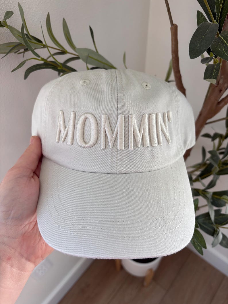 May include: A light gray baseball cap with the word "MOMMIN'" embroidered in a lighter shade of gray across the front. The cap has a curved brim and a classic six-panel design. The background includes greenery.