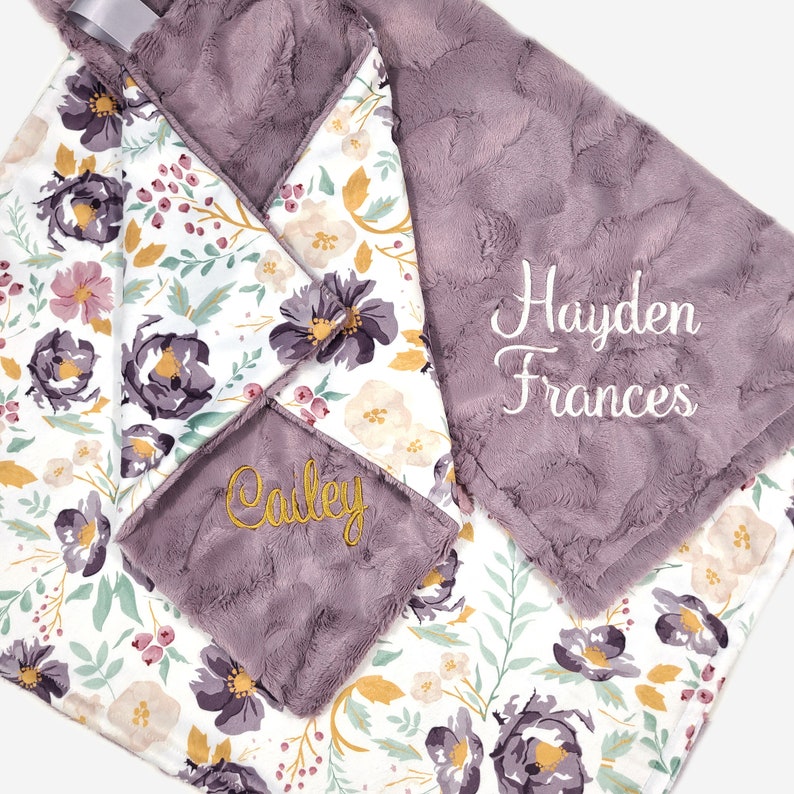 Puede incluir: Una manta de beb&eacute; floral de color morado y blanco con los nombres "Hayden Frances" y "Cailey" bordados en hilo dorado.