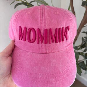 May include: A pink baseball cap with the word "MOMMIN'" embroidered across the front in a darker pink. The cap is made of a soft fabric and has a curved brim. The hat is a solid pink color.