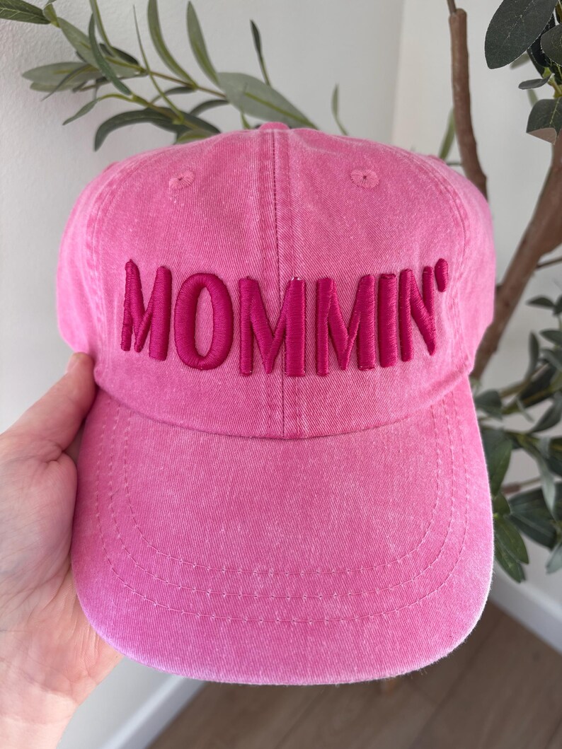 May include: A pink baseball cap with the word "MOMMIN'" embroidered across the front in a darker pink. The cap is made of a soft fabric and has a curved brim. The hat is a solid pink color.
