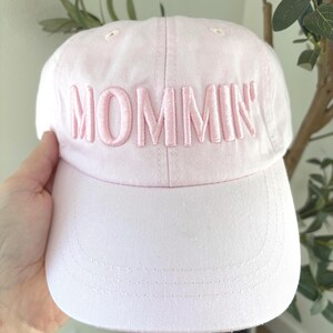 May include: A light pink baseball cap with the word "MOMMIN'" embroidered in a matching pink thread across the front. The cap has a curved brim and a classic six-panel design. A casual accessory.