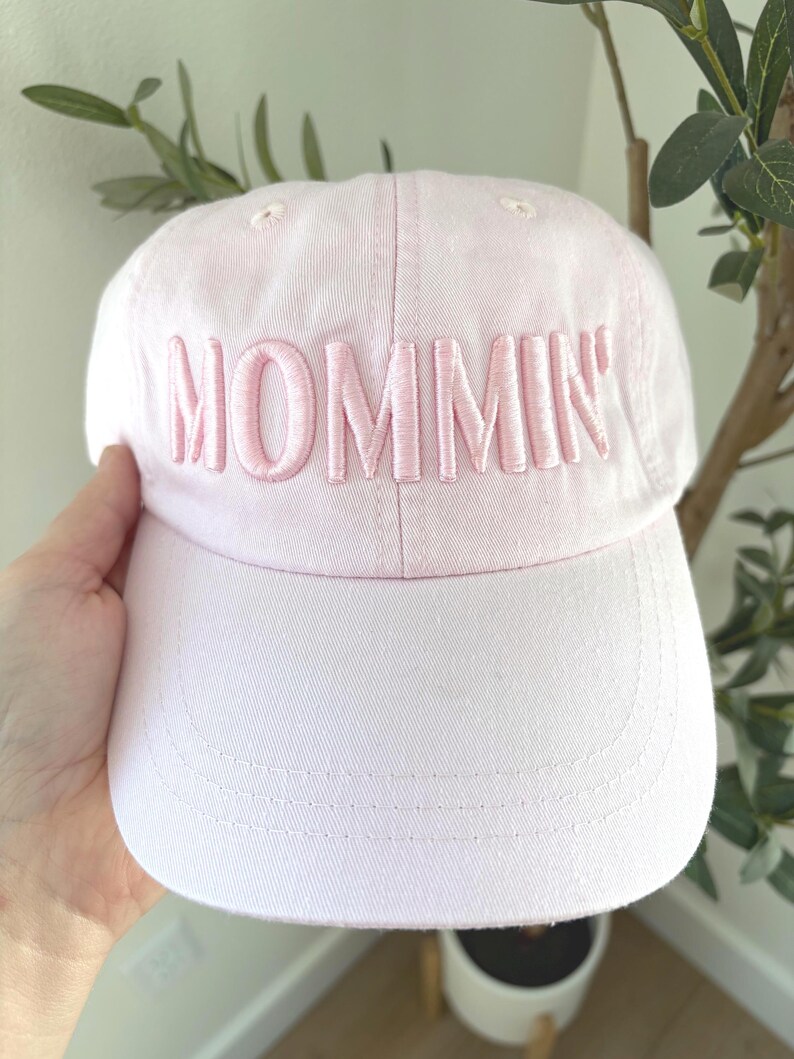 May include: A light pink baseball cap with the word "MOMMIN'" embroidered in a matching pink thread across the front. The cap has a curved brim and a classic six-panel design. A casual accessory.