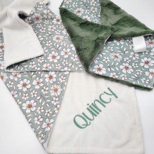 Peut inclure: Une couverture pour b&eacute;b&eacute; verte et blanche avec un motif floral. La couverture est personnalis&eacute;e avec le nom "Quincy" brod&eacute; en fil vert.