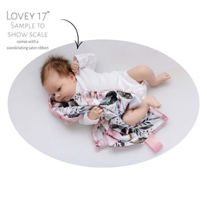 May include: A baby is lying on a white surface with a pink and white floral patterned baby blanket. The blanket has a pink satin trim. The text "LOVEY 17" is visible in the image.
