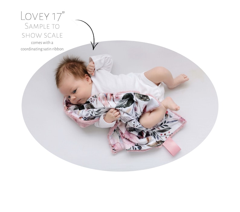 May include: A baby is lying on a white surface with a pink and white floral patterned baby blanket. The blanket has a pink satin trim. The text "LOVEY 17" is visible in the image.