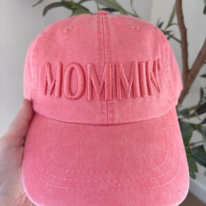 May include: A pink baseball cap with the word "MOMMIN" embroidered across the front. The cap has a curved brim and a classic six-panel design. The hat is made of a soft, textured fabric.