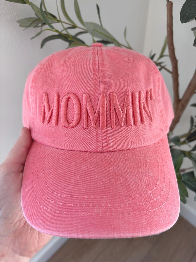 May include: A pink baseball cap with the word "MOMMIN" embroidered across the front. The cap has a curved brim and a classic six-panel design. The hat is made of a soft, textured fabric.