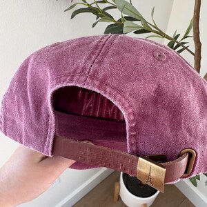May include: A dusty rose baseball cap with a curved brim. The adjustable strap is brown leather with embossed text and a gold-colored metal buckle featuring the Eiffel Tower. The cap is made of a textured fabric.