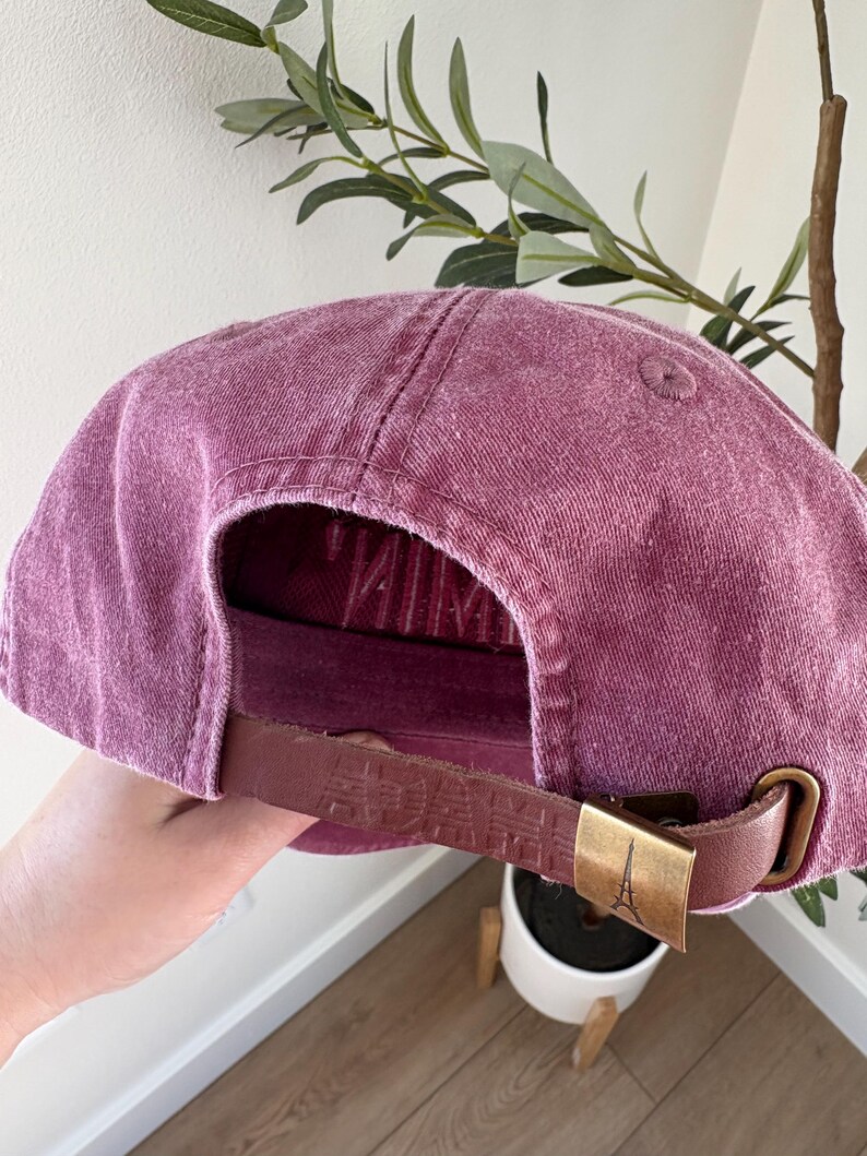 May include: A dusty rose baseball cap with a curved brim. The adjustable strap is brown leather with embossed text and a gold-colored metal buckle featuring the Eiffel Tower. The cap is made of a textured fabric.