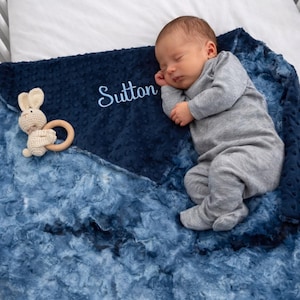 May include: A sleeping infant in a gray onesie rests on a navy blue and blue tie-dye patterned blanket. The blanket is embroidered with the name "Sutton" in light blue. A small, beige bunny-shaped teether toy is on the blanket.