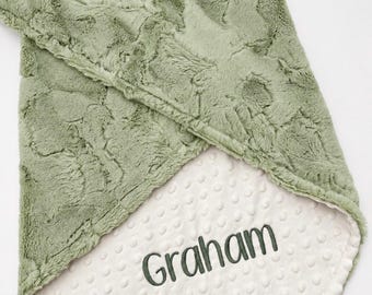 May include: A sage green faux fur blanket with a textured, plush surface. The underside is a cream-coloured minky fabric featuring a raised dot pattern. The name Graham is embroidered in dark green thread on the cream-coloured side of the blanket.