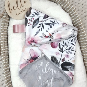 May include: A gray and white baby blanket with a floral print and the name "Alora Kent" embroidered on it. The blanket is in a wicker basket with a wooden sign that says "hello world".