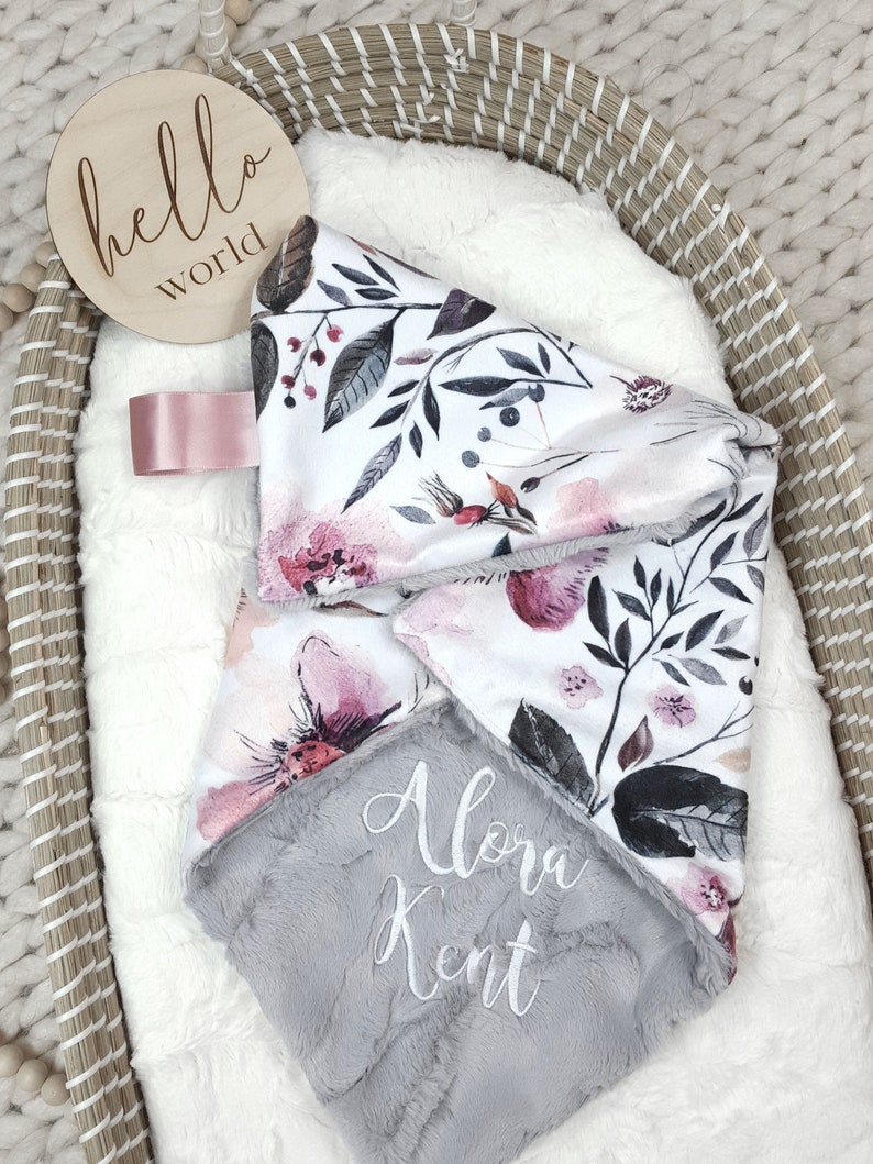 May include: A gray and white baby blanket with a floral print and the name "Alora Kent" embroidered on it. The blanket is in a wicker basket with a wooden sign that says "hello world".