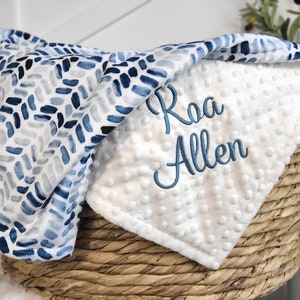 May include: A white and blue baby blanket with a blue and white watercolor pattern on one side and a white minky fabric on the other side. The blanket is personalized with the name "Raa Allen" embroidered in blue thread.