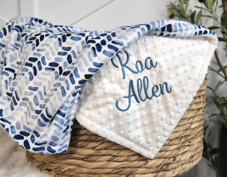 May include: A white and blue baby blanket with a blue and white watercolor pattern on one side and a white minky fabric on the other side. The blanket is personalized with the name "Raa Allen" embroidered in blue thread.