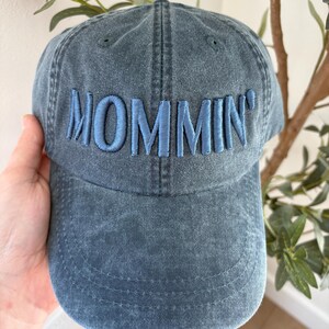 May include: A denim blue baseball cap with the word "MOMMIN'" embroidered in a lighter blue thread across the front. The cap has a curved brim and a classic six-panel design, perfect for casual wear.