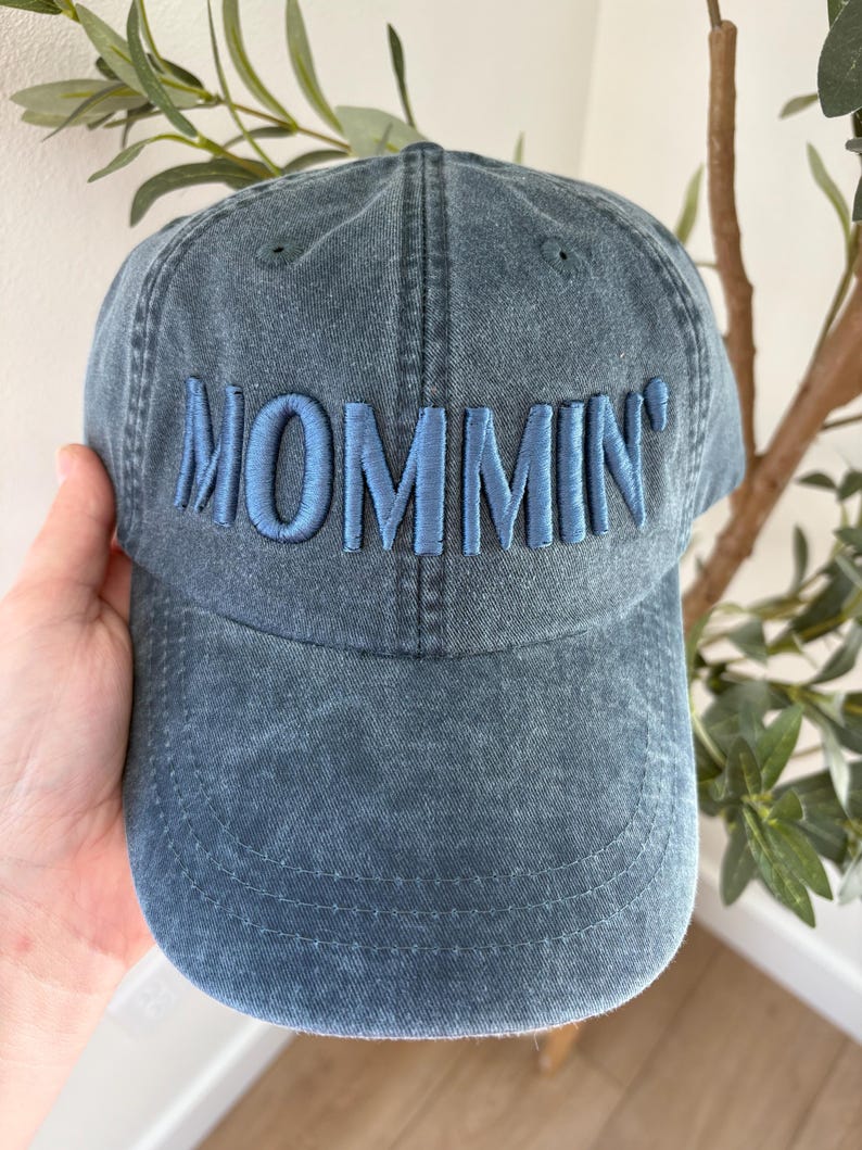 May include: A denim blue baseball cap with the word "MOMMIN'" embroidered in a lighter blue thread across the front. The cap has a curved brim and a classic six-panel design, perfect for casual wear.