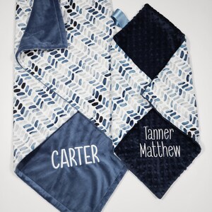 May include: Two blue and white baby blankets with personalized embroidery. One blanket features a chevron pattern and the name "CARTER" embroidered on the blue side. The other blanket features a navy blue side with the names "Tanner Matthew" embroidered on it.