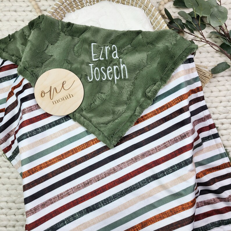 May include: A green and white striped baby blanket with a plush green minky side. The blanket has the name "Ezra Joseph" embroidered on it. A wooden circle with the words "one month" is on top of the blanket.