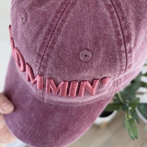 May include: A dusty rose baseball cap with the word "Commin'" embroidered on the front. The cap has a curved brim and a stitched design. The hat is made of a soft, textured fabric.