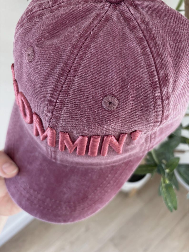 May include: A dusty rose baseball cap with the word "Commin'" embroidered on the front. The cap has a curved brim and a stitched design. The hat is made of a soft, textured fabric.
