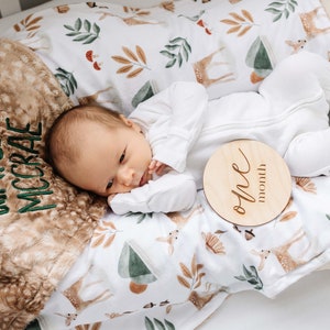May include: A baby lying on a white and brown blanket with a deer and leaf pattern. The baby is wearing a white onesie and is holding a wooden sign that says "one month". The blanket has the name "Waylon McCrae" embroidered on it in green thread.