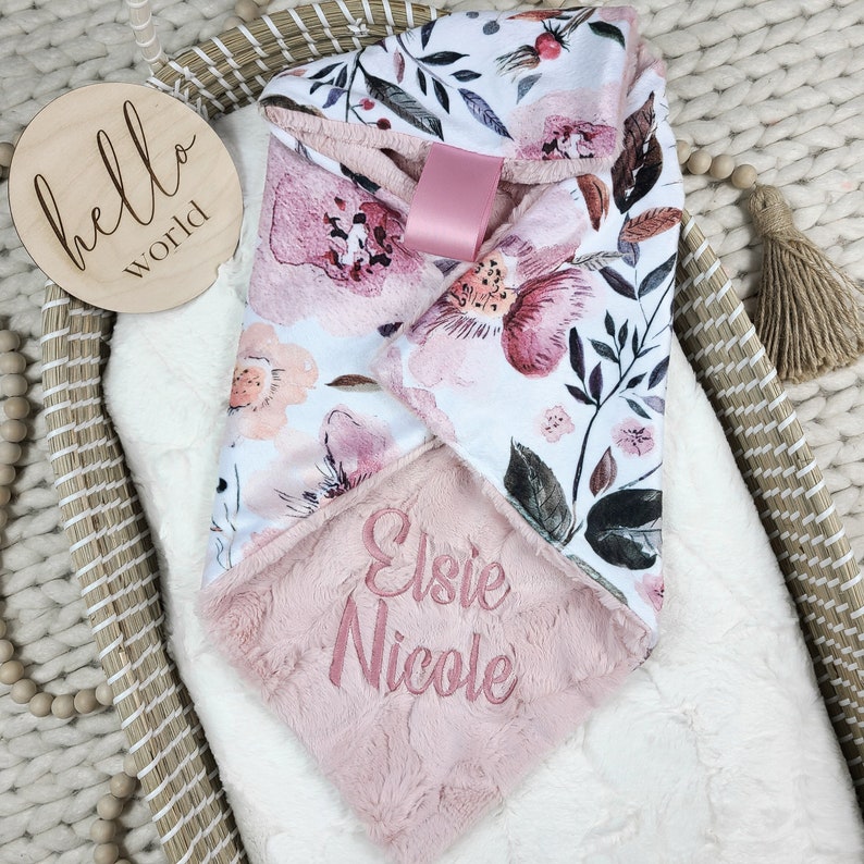 May include: A pink and white floral print baby blanket with a soft pink minky backing. The blanket is personalized with the name "Elsie Nicole" embroidered in pink thread.