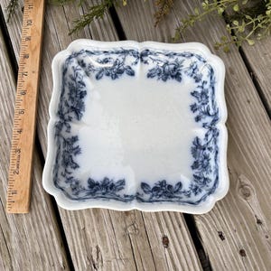 May include: A square white ceramic dish with scalloped edges and a blue floral pattern along the border. A wooden ruler is placed next to the dish, showing it measures approximately 6 inches in width and length.