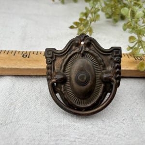 May include: Two antique bronze-colored drawer pulls with a decorative design. Each pull features an oval center with a detailed border and a curved handle. A wooden ruler is visible in the background, along with some greenery.