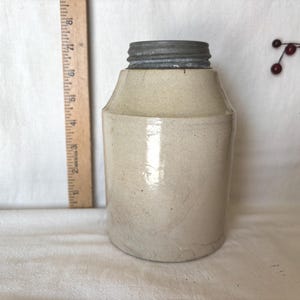 Puede incluir: Un tarro de gres antiguo con tapa de metal. El tarro es de color blanquecino con una superficie ligeramente texturizada y forma cilíndrica. Una regla de madera está colocada junto al tarro. El tarro mide unos 18 cm de alto.