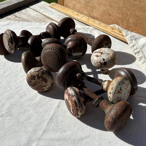 Puede incluir: Una colección de pomos de puerta vintage con varios acabados, incluyendo metal marrón oxidado y superficies pintadas de blanco desgastado con desgaste visible. Los pomos están agrupados, algunos con bordes de cuentas decorativos, junto a una regla de madera.