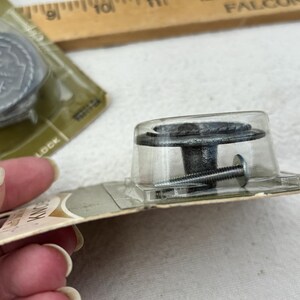 Puede incluir: Pomos de armario de metal envejecido con un diseño decorativo, empaquetados en plástico transparente. Los pomos tienen un acabado oscuro y vienen con tornillos de montaje. El embalaje está sobre una superficie blanca.
