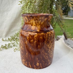 May include: A cylindrical ceramic crock featuring a glossy, mottled brown and tan glaze. The vessel has a slightly flared rim and a smooth, rounded body, showcasing a distinct drip-style pattern throughout the exterior surface.