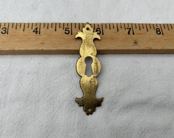 Tapa de latón para cerradura, forma oblonga con parte superior acampanada y clavos de fijación inferiores incluidos.