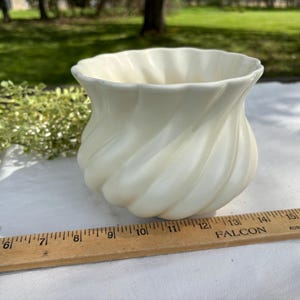 May include: Cream-colored ceramic vase with a spiral design. The vase has a wide opening and a rounded base. A wooden ruler with inch measurements is in the foreground. The vase is sitting on a white surface.