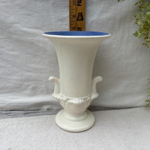 May include: A white ceramic vase with a flared rim and a blue interior. The vase has two curved handles and a decorative base. The vase is approximately 23 cm tall.