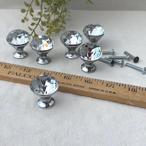 May include: A collection of seven crystal cabinet knobs with silver-colored bases and mounting screws. The knobs are faceted, reflecting light. A wooden ruler with inch markings is also present, along with some greenery.
