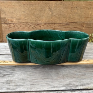 May include: A dark green ceramic planter with a smooth, glossy finish and a three-lobed, clover-like shape. The planter is positioned behind a wooden ruler, showing a length spanning from the 11-inch mark to the 27-inch mark on the scale.