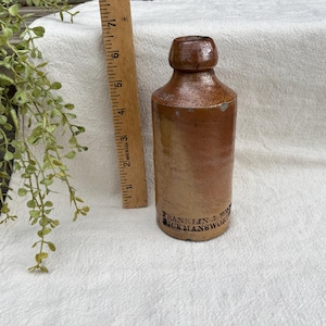 Puede incluir: Una botella de gres marrón antiguo con una parte superior redondeada y un cuerpo cilíndrico. La botella tiene una etiqueta descolorida con el texto "FRANKLIN & SON, HICKMANSWORTH". Una regla de madera está al lado de la botella. La botella mide aproximadamente 18 cm de alto.