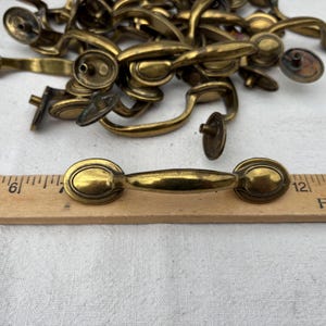 Op de afbeelding: Een verzameling antieke messing lade-knoppen. De knoppen hebben een ovale achterplaat en een gebogen handvat. Een houten liniaal met centimeter markeringen staat op de voorgrond. De knoppen hebben een warme, verouderde messing kleur.