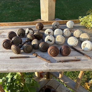 May include: A collection of round door knobs in various finishes, including dark brown metal and distressed white paint. Several knobs feature attached metal spindles. A wooden ruler is visible at the top, showing measurements in inches.