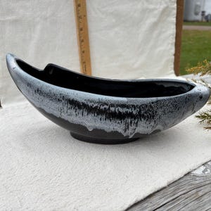 Peut inclure: Un bol en céramique noir et blanc de forme allongée. L'intérieur du bol est noir brillant et l'extérieur est texturé, blanc et noir. Le bol mesure environ 30 cm de long et 15 cm de large, avec une base basse et arrondie.