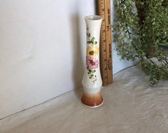 May include: A white ceramic vase with a floral design featuring pink, yellow, and green flowers. The vase has a brown base and a slender, cylindrical shape.