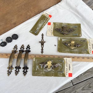 May include: An assortment of vintage brass hardware, including drop pulls and decorative handles, arranged on a white cloth. The hardware is in various shapes and sizes, with a ruler for scale. Some pieces are still in their original packaging.