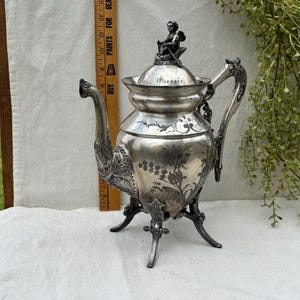 May include: An antique silver teapot with ornate floral and scrollwork designs. The teapot has a decorative handle, a curved spout, and a small cherub figure on the lid. It stands on three legs and is displayed on a white surface.