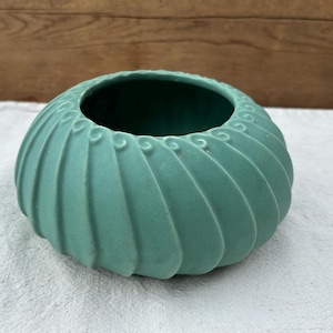 May include: A matte seafoam green ceramic bowl featuring a rounded, low-profile shape with a series of vertical, swirling fluted ridges that spiral toward the center opening. The interior of the bowl is a darker, solid green finish.