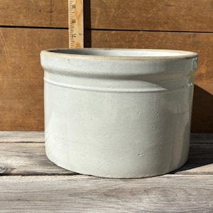 May include: A cylindrical off-white stoneware crock with a smooth glazed finish and a thick rim. A wooden ruler is positioned behind the vessel, showing a height measurement of approximately 7 inches from the base to the top edge of the rim.