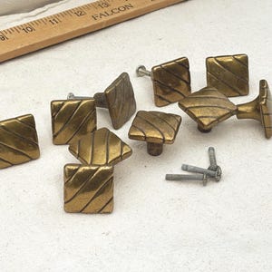 May include: A collection of eight square brass cabinet knobs featuring diagonal grooved lines, accompanied by three silver-colored metal screws. A wooden ruler is visible at the top, showing measurements in inches for scale reference.