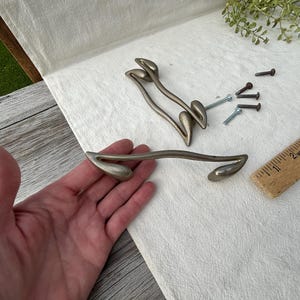 Op de afbeelding: Een set kastgrepen in antieke stijl met een zilveren afwerking. De grepen hebben een gebogen, organisch ontwerp. Ook inbegrepen zijn schroeven en een houten liniaal. De grepen zijn ongeveer 13 cm lang.
