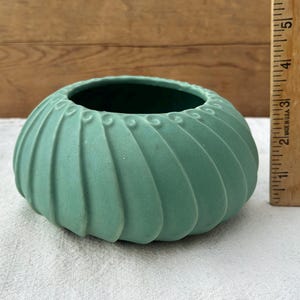 May include: A matte sage green ceramic bowl with a ribbed, swirling exterior pattern and a wide circular opening. A wooden ruler is placed vertically next to the bowl, showing a height of approximately 2.5 inches for the object.